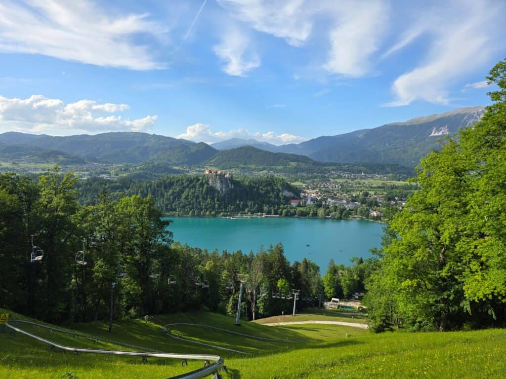 View of Lake Bled from Straza Hill at the top of chair lift and alpine side with blue sky and mountains in the distance and the grassy downhill in front.