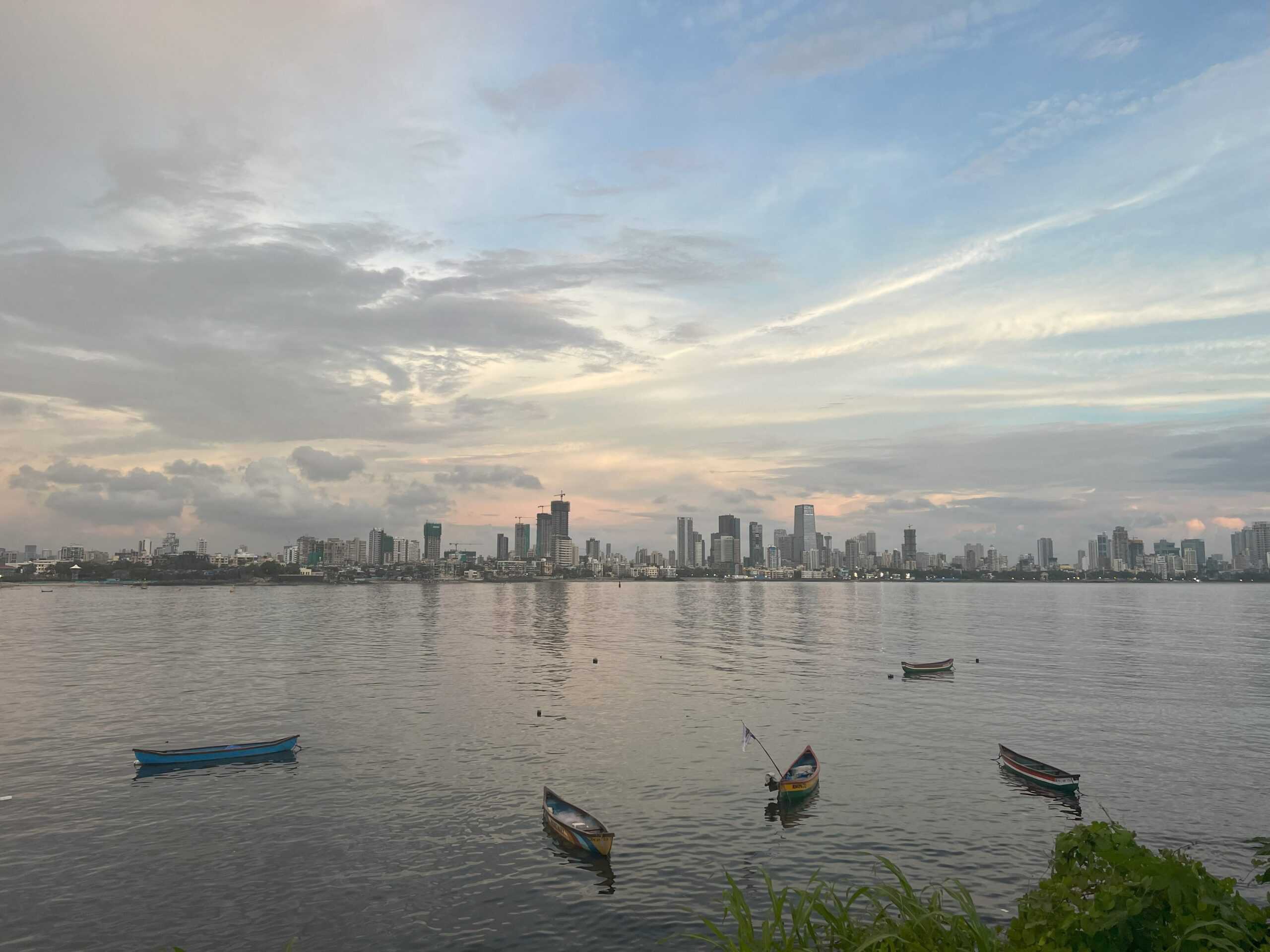 A sunset view from Bandra Reclamation in Mumbai, overlooking calm waters with several small boats floating idly. The skyline is dotted with the silhouettes of buildings, under a sky painted with soft hues of blue, pink, and white clouds.