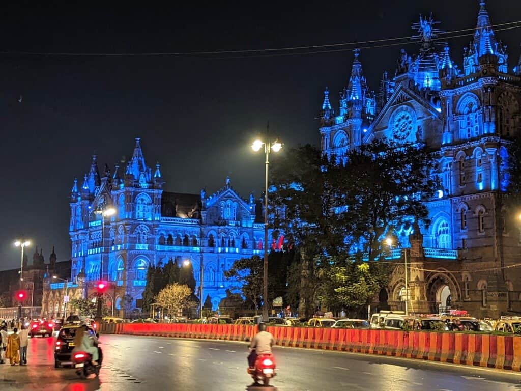 Nighttime view of the illuminated Chhatrapati Shivaji Terminus in Mumbai, showcasing its intricate Gothic architecture with vibrant blue lighting. The street in front reflects the city's dynamic energy with moving vehicles and pedestrians.