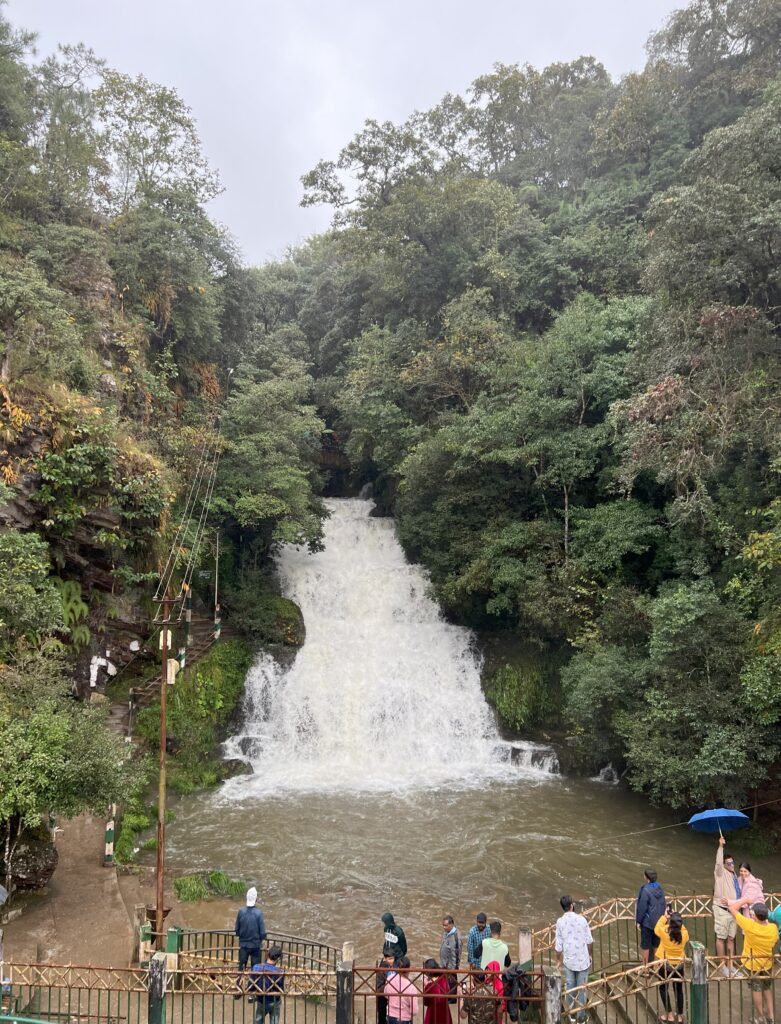 View of Elephant Falls in Shillong from across the river with people standing on the walkway to admire the waterfall.