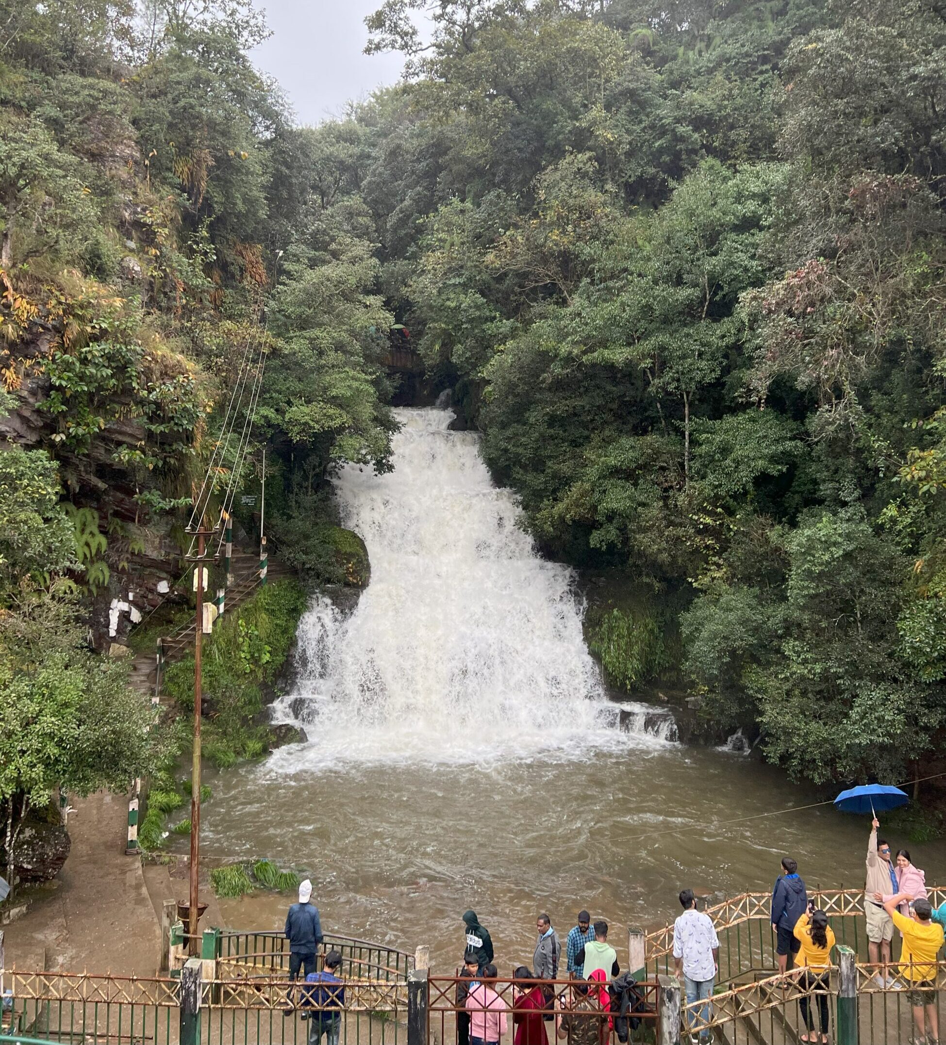 View of Elephant Falls in Shillong from across the river with people standing on the walkway to admire the waterfall.