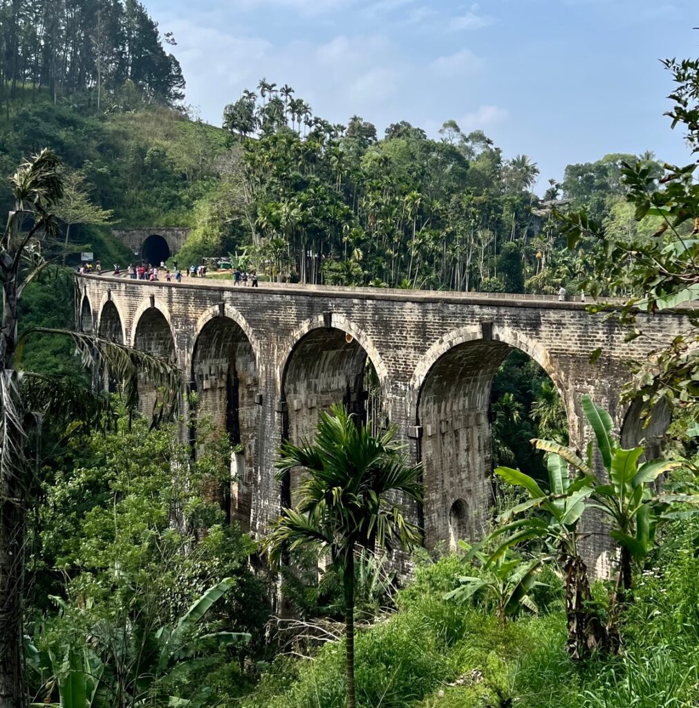 Nine Arches Bridge spanning across a lush tropical valley. The bridge is made of weathered gray stone or brick and features classic colonial architecture. People can be seen walking along the top of the bridge, and there's a tunnel entrance visible at one end. The surrounding landscape is filled with palm trees, banana plants, and dense tropical vegetation. 