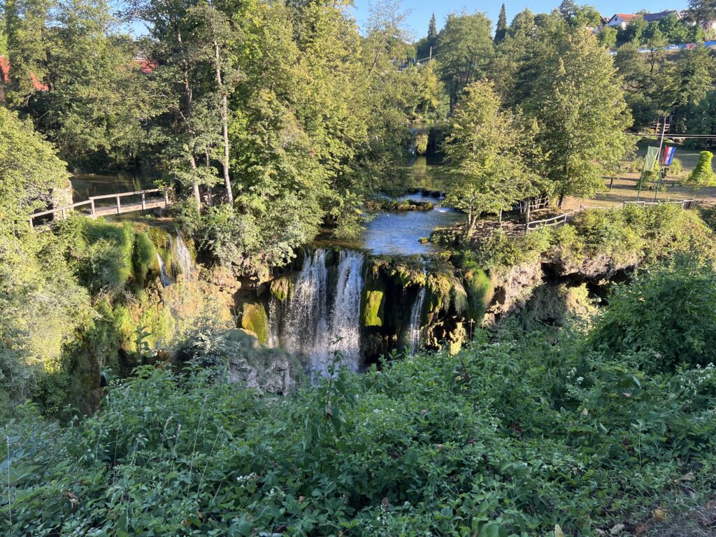 Trees, a wooden bridge, and a small waterfall along a pathway in Rastoke, Croatia