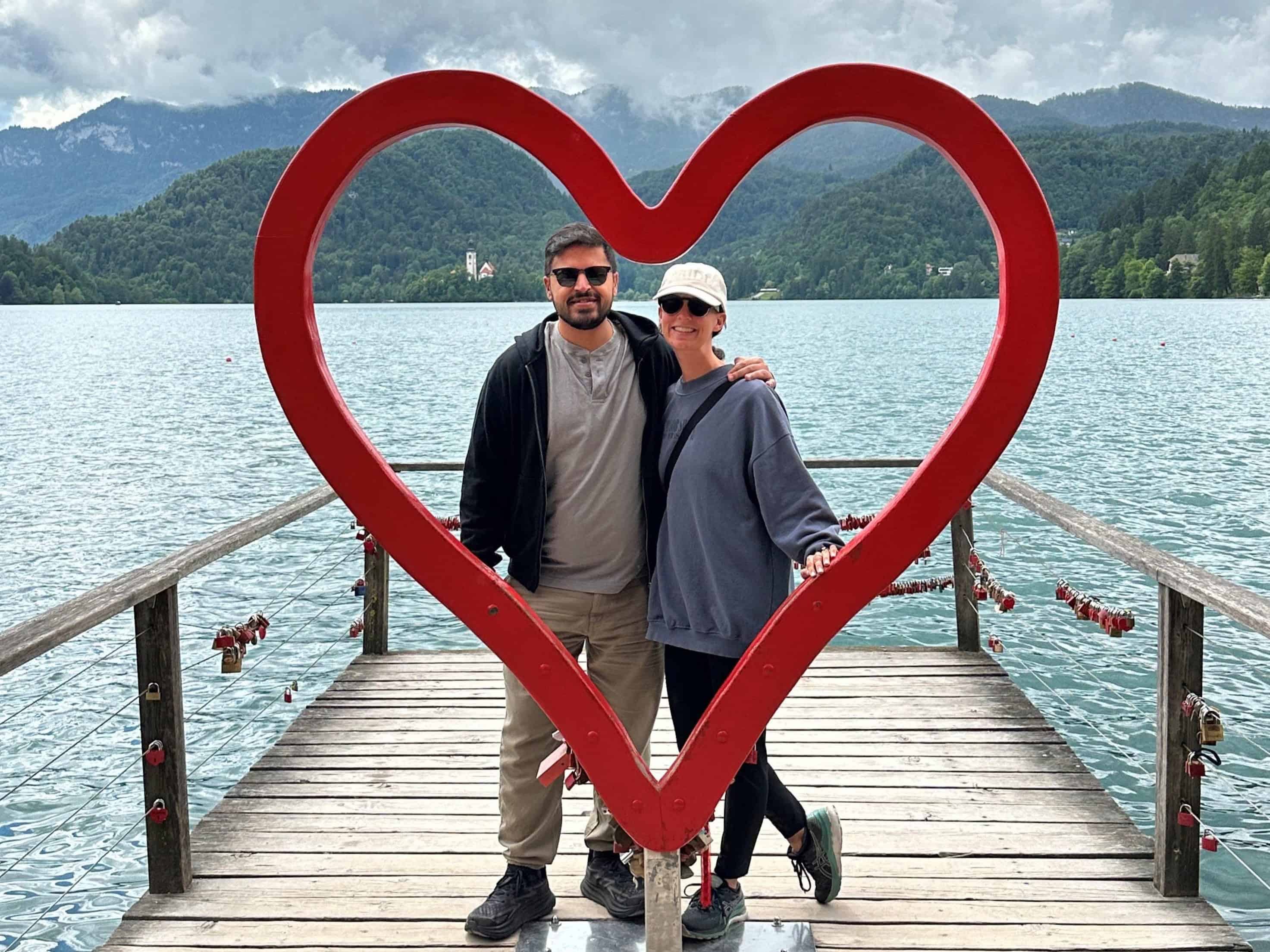 Couple standing in the large cut out heart frame on a pier on Lake Bled with the water and mountains in the background.