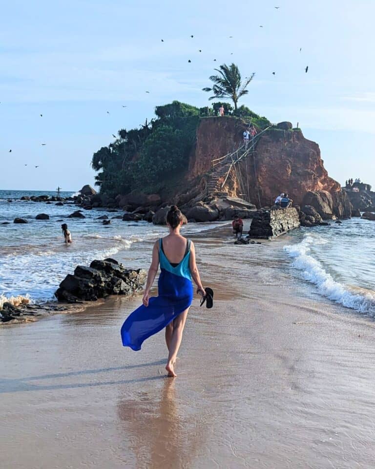 The view from behind a girl walking barefoot in a blue dress towards Parrot Rock on the beach in Mirissa.