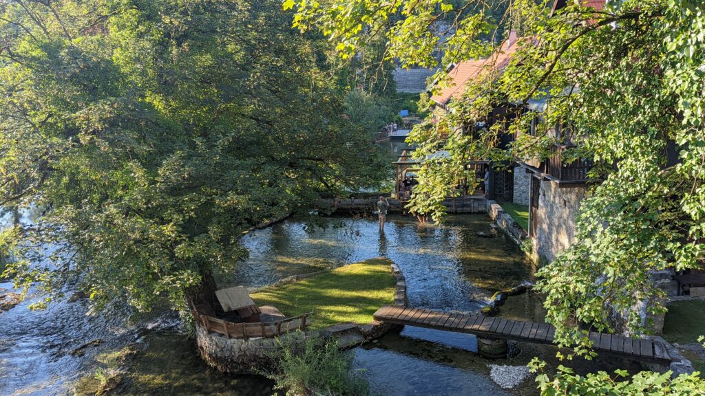 View of the river and a small patch of grass surrounded by water and connected with a small wooden bridge in the backyard of a local's house in Rastoke, Croatia