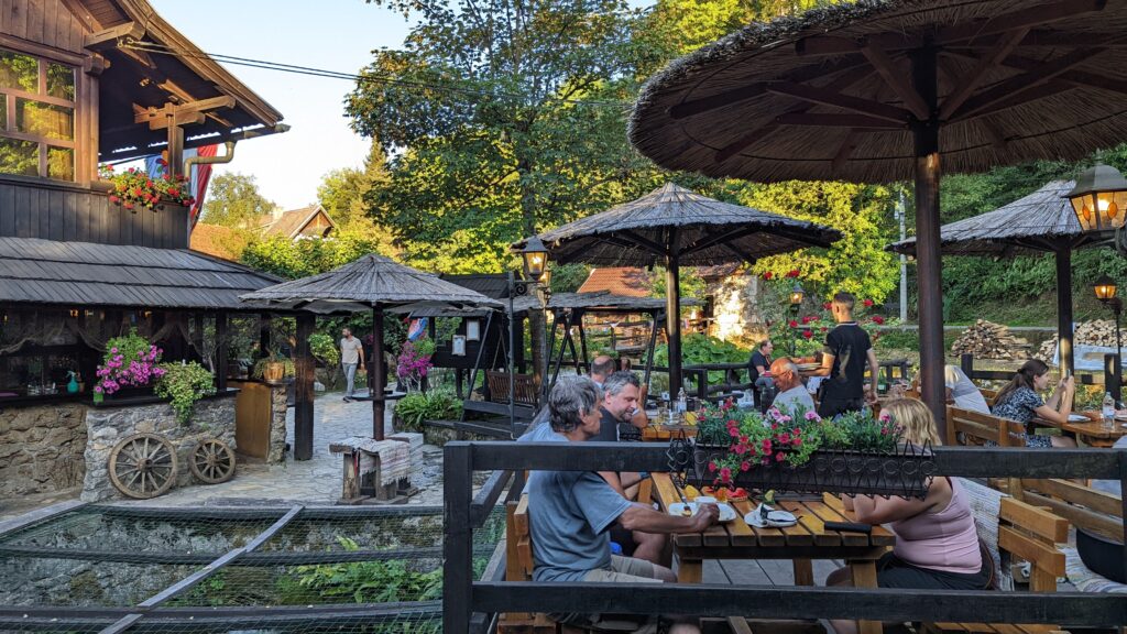 People eating in a quaint setting with wooden tables and straw sun umbrellas at Petro Restaurant in Rastoke, Croatia.