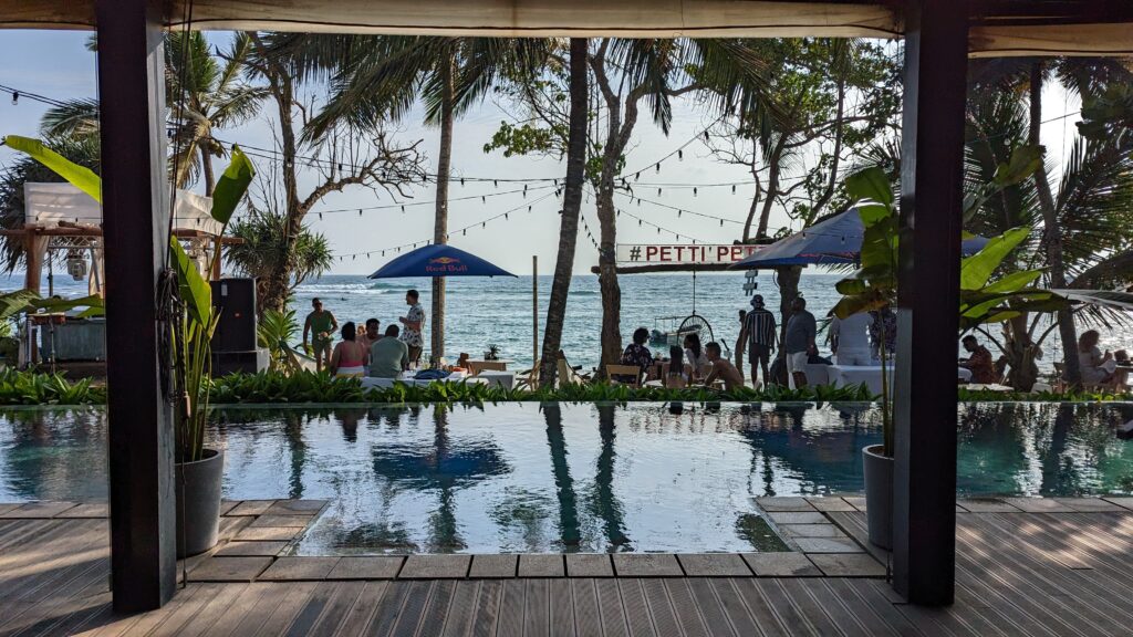 A view of Petti Petti, a beachside cafe and bar in Mirissa, showing a pool with tables and chairs beyond it and the ocean in the distance.