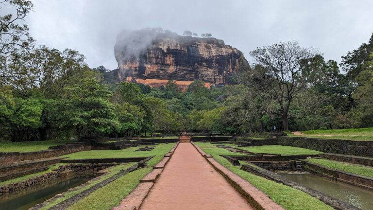 A wide pathway leading through lush gardens toward Sigiriya Rock, an ancient fortress in Sri Lanka. The massive rock formation is partially covered by mist, with greenery and ruins surrounding the area.