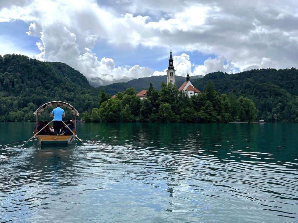 View of the back of a wooden Pletna boat being rowed to Bled Island in Lake Bled with a cloudy sky and mountains in the background of the turquoise lake.