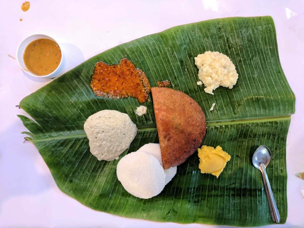 A traditional South Indian meal is served on a large banana leaf. The leaf holds a variety of dishes, including two soft idlis (steamed rice cakes), a crispy half dosa (savory crepe), a dollop of yellow ghee, a serving of white rice pudding, and a side of chutneys and sambar. A cup of sambar sits to the side of the leaf, and a spoon rests on the right edge.