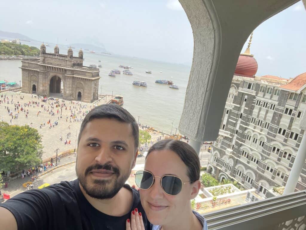 Two people taking a selfie on a balcony from Taj Mahal Palace hotel with the Gateway of India down below in the background and the sea behind that.