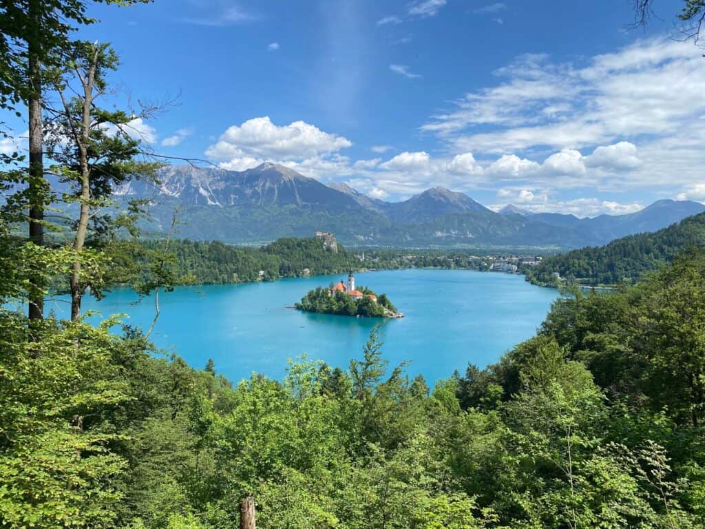 View of Lake Bled with Bled Island in the middle from the Mala Osojnica hike. Blue skies in the background and green mountains surrounding the turquoise lake.