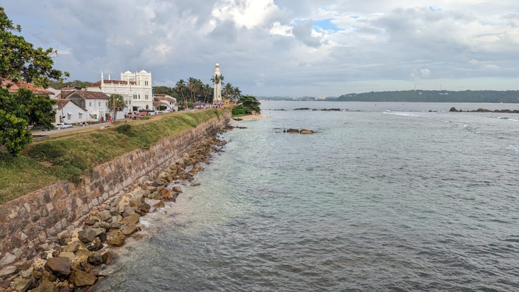 A view of the Galle Fort coastline in Sri Lanka, with a stone wall lining the shore, historic white buildings, and a lighthouse surrounded by palm trees. The calm ocean stretches out under a partly cloudy sky.