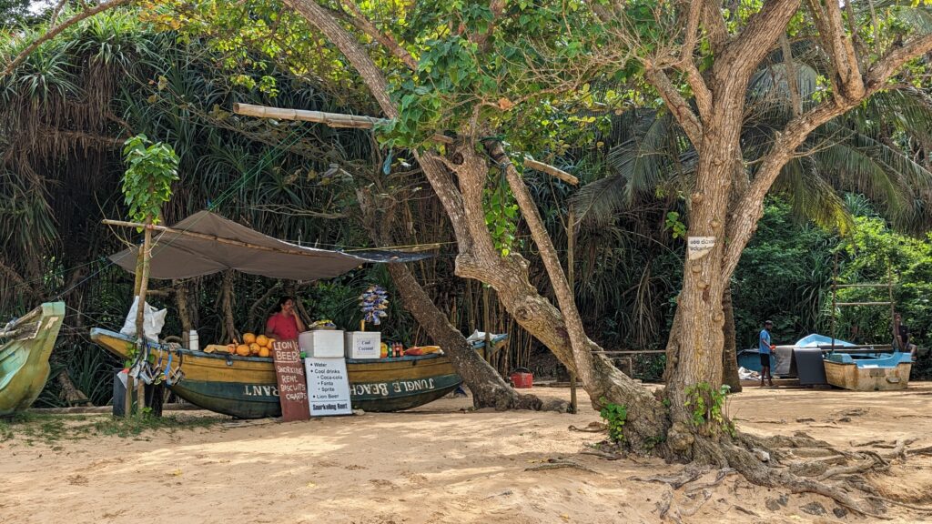 Small refreshment stand set up in a repurposed fishing boat under a shaded tree at Jungle Beach, Unawatuna, Sri Lanka. A vendor stands by the stall offering coconuts, water, cold drinks, and snacks against a backdrop of lush tropical greenery.