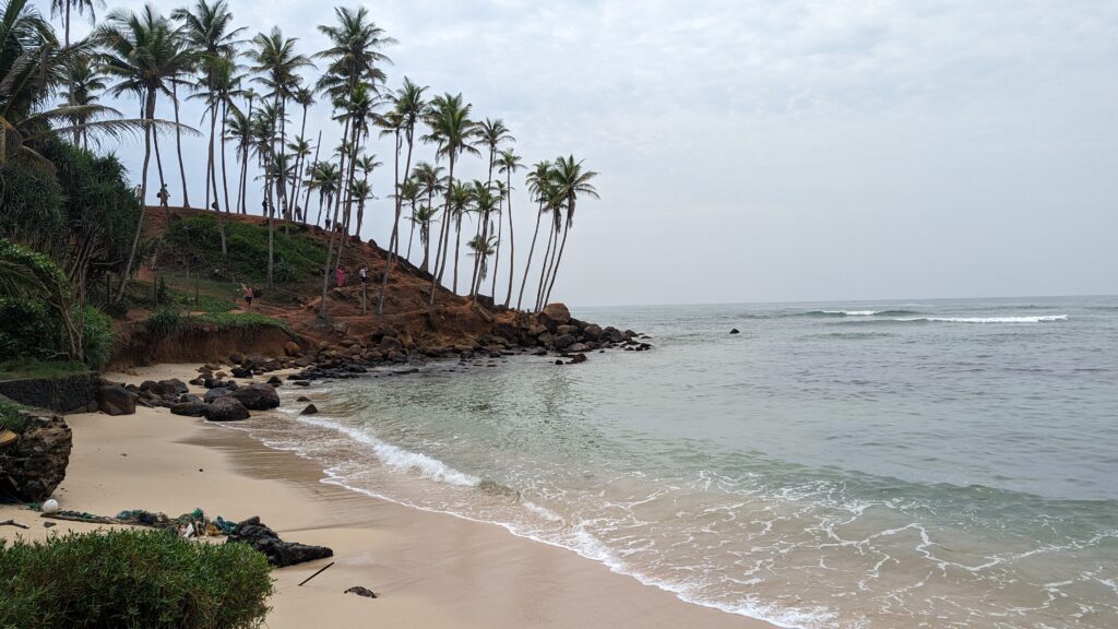 A beach with small waves, sand, and a small rocky area at the shoreline. A picturesque hill covered with tall, slender palm trees leans toward the ocean under a cloudy sky, creating a tropical, idyllic scene at Coconut Tree Hill in Mirissa, Sri Lanka.