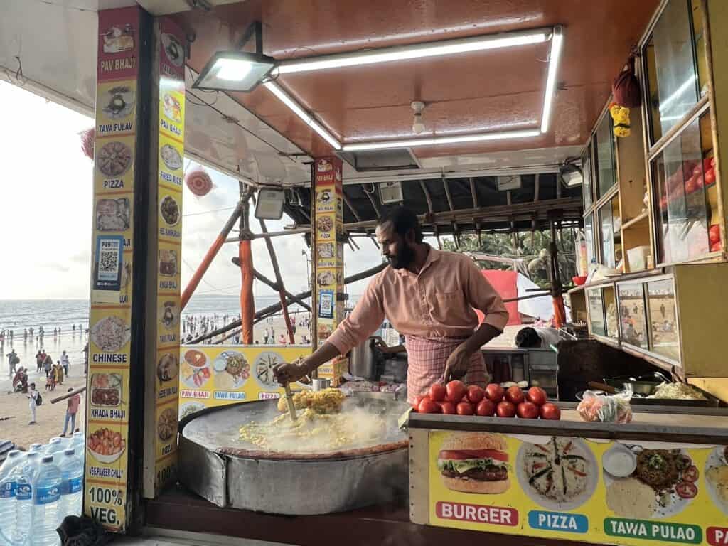 A food vendor is preparing pav bhaji on a large flat griddle at a beachside stall. Menus with various food options, including Chinese and pizza, are displayed around the stall. The scene is set against the backdrop of Juhu Beach in Mumbai, with people and the ocean visible in the distance