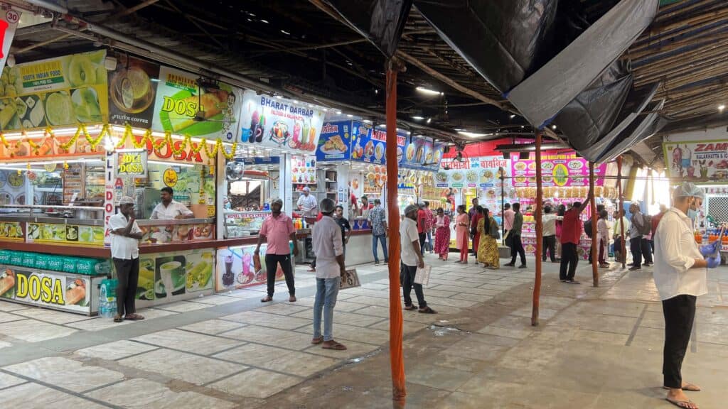 A food court with multiple street food stalls at Juhu Beach, Mumbai. The stalls are brightly lit and display colorful signs for South Indian food like dosa and idli, as well as other snacks. People are seen ordering food and walking around the area, which covered with a tin roof.