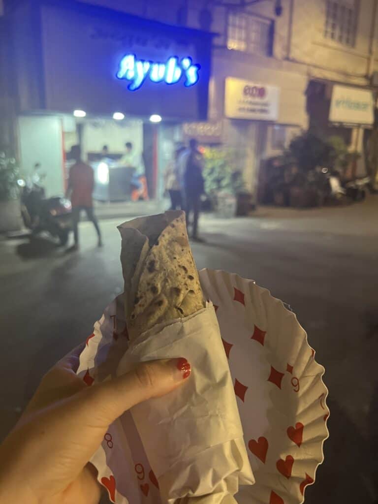 A close-up of a person's hand holding a wrapped kebab. In the background, a neon-lit sign reads 'Ayub's' on an urban street at night, with people and a scooter visible in the distance.