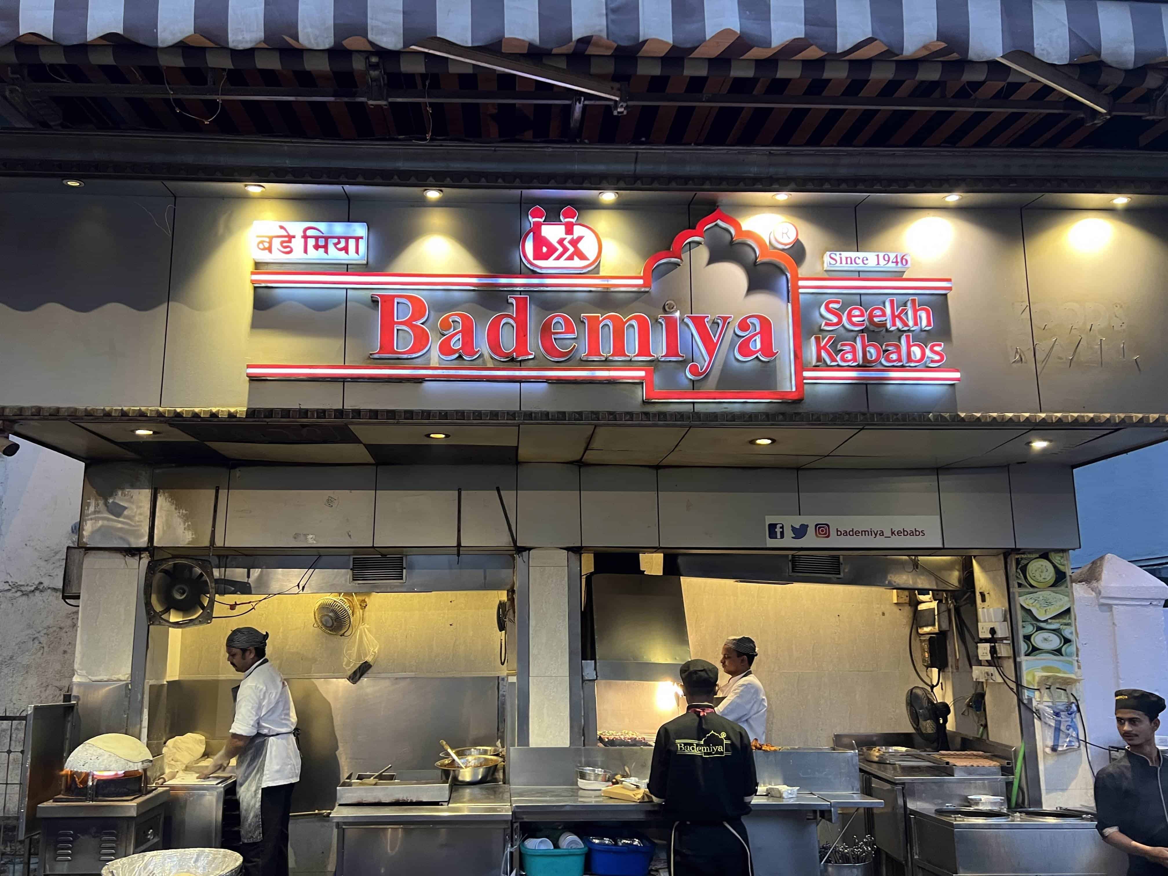 A street food stall named 'Bademiya Seekh Kababs' with a vibrant red and white signboard that includes a crown symbol and the text 'Since 1946'. The stall has an open front with two chefs working inside, one managing a grill and the other preparing food. The stall is equipped with stainless steel kitchen equipment and has fans on the wall. 