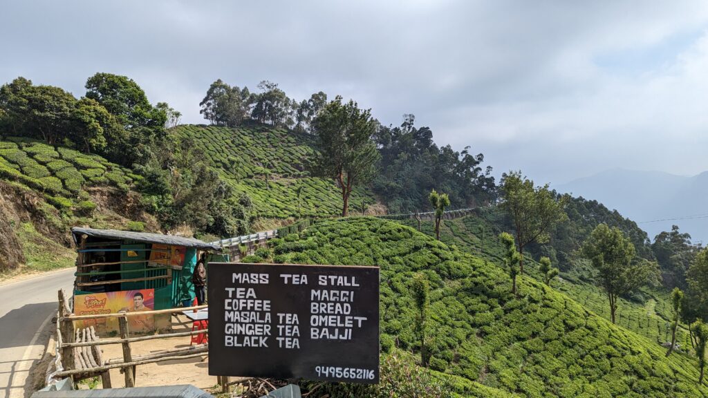 Roadside tea stand with sign advertising its menu and rolling green hills with tea plantations in the background.