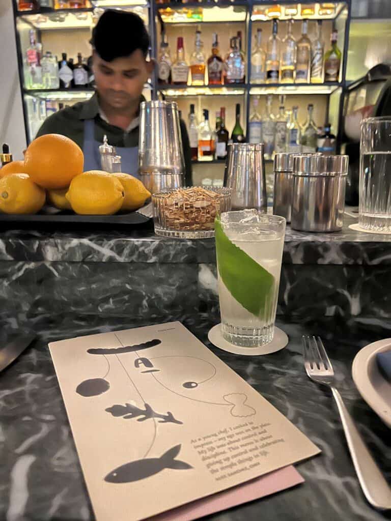 Americano bar with shelves of liquor bottles in background and a light green cocktail and menu on the bar in the foreground.