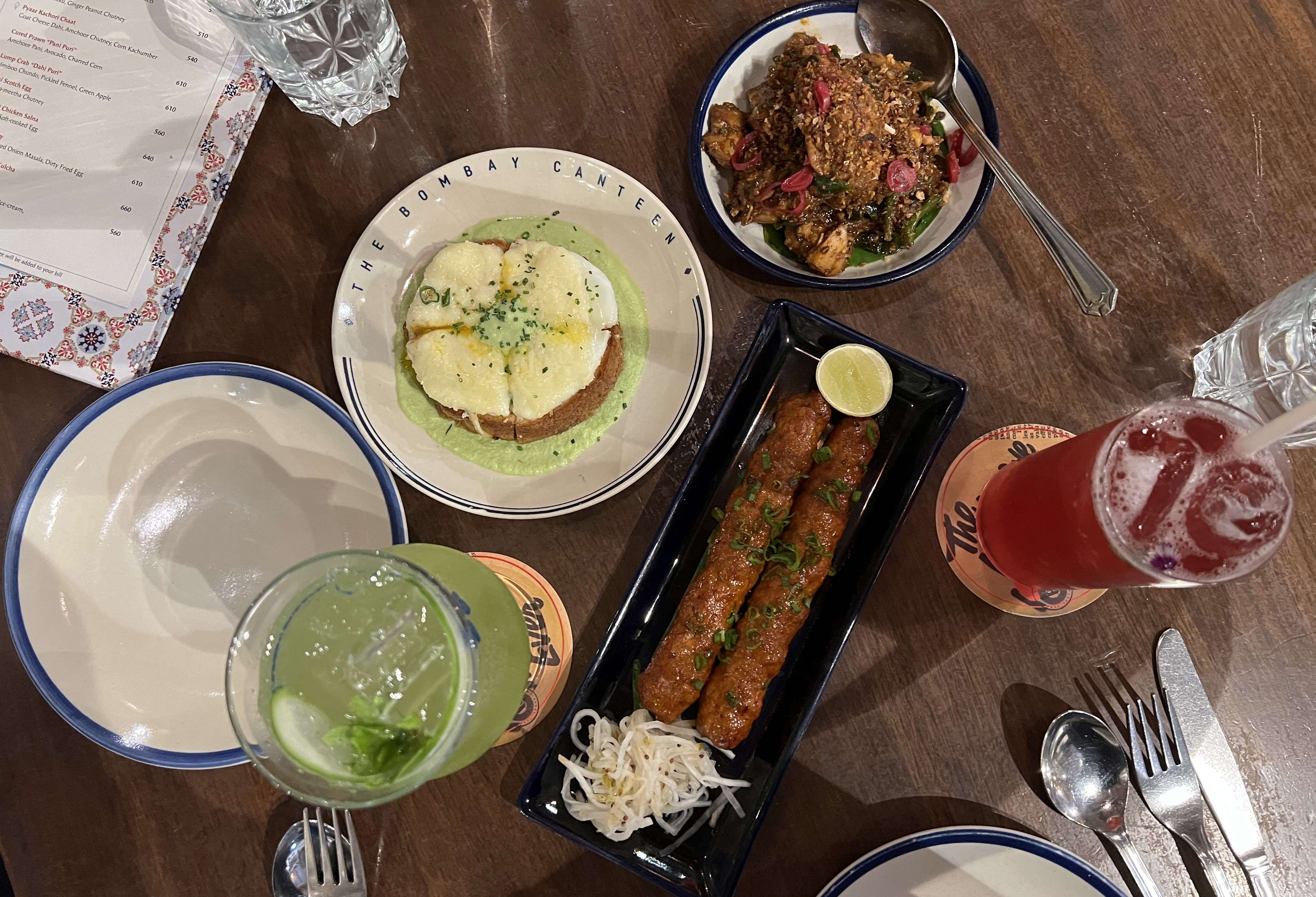 Overhead view of table at Bombay Canteen in South Mumbai with three plates of food, a couple cocktails and a corner of the menu.