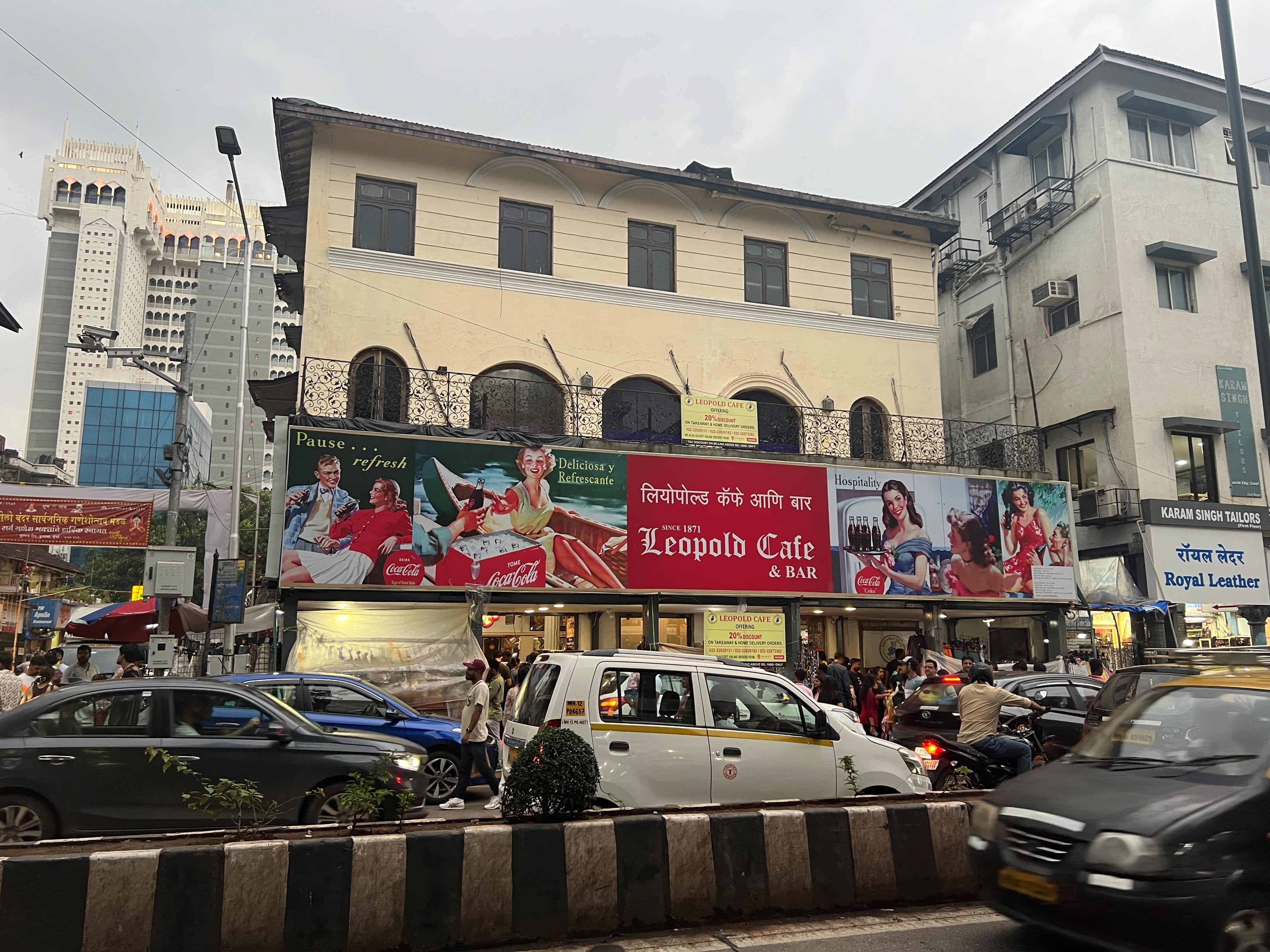 The outside of Cafe Leopold in Mumbai with the busy street and cars in front of it.