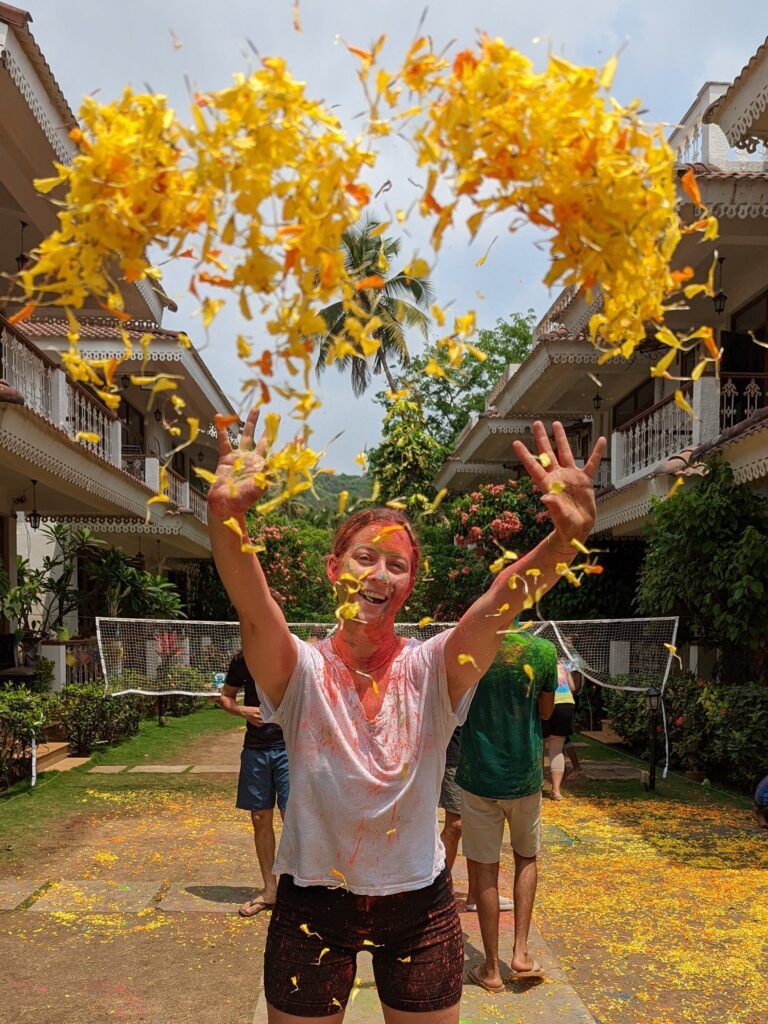 A girl covered in bright colored powder throwing flowers in the air to celebrate Holi in India.