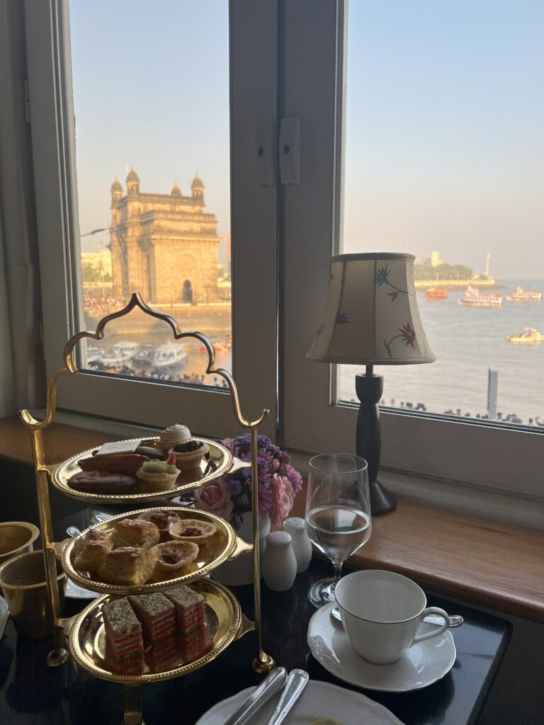 A three tiered tray of tea time snacks and a tea cup on a table by a window at the Sea Lounge at Taj Mahal Palace hotel in South Mumbai. Out of the window you can see the Gateway of India in the background.