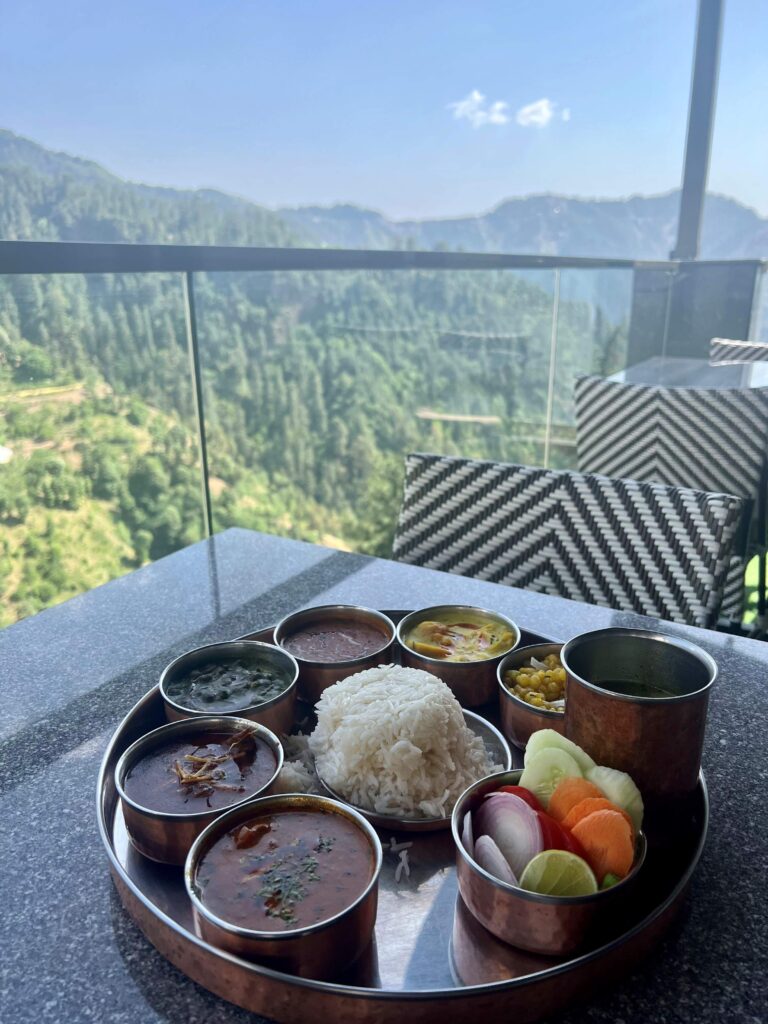 Himachali Thali with rice in the middle and different small dishes surrounding it on a table with the Himalayas in the background.