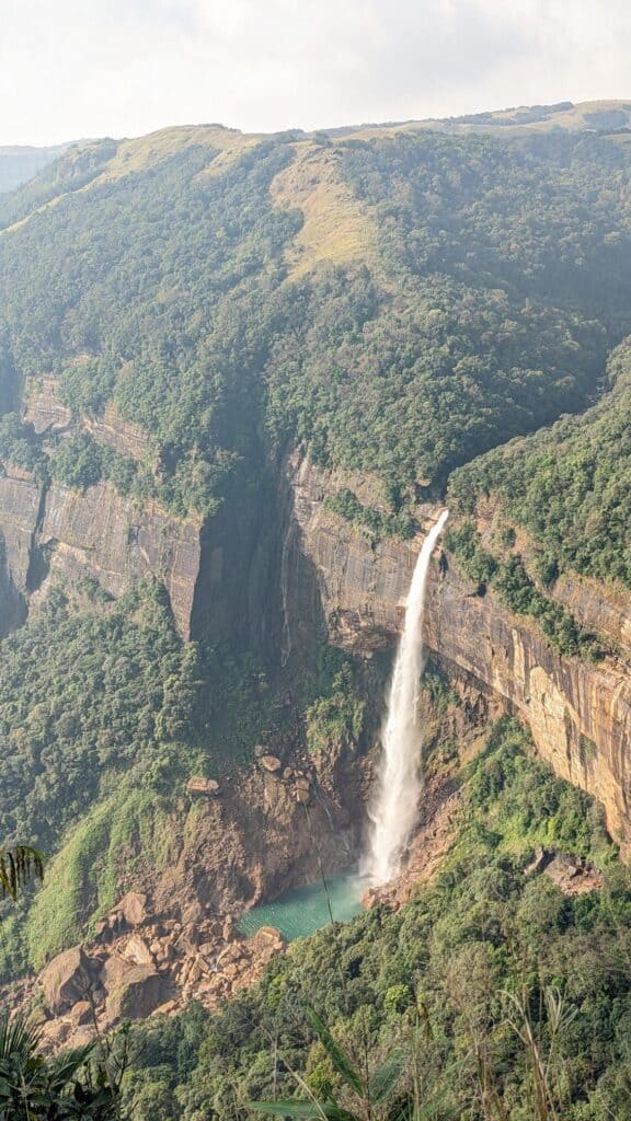 View of Nohkalikai Waterfall, plunging off the lush green cliffside into a blue water pool.