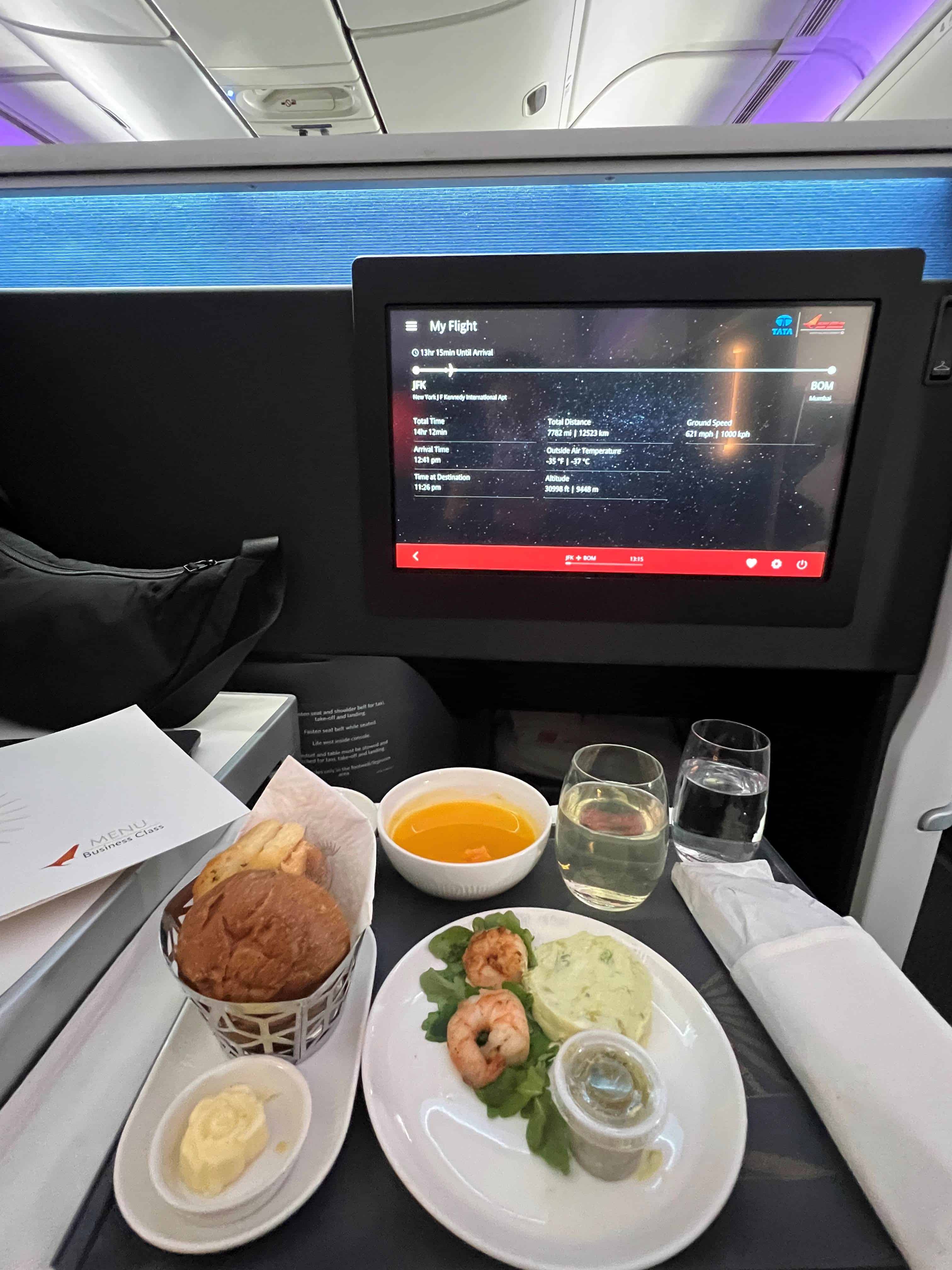 Air India airplane dinner appetizers including shrimp, soup, and bread plated on a tray table with a video screen in the background.