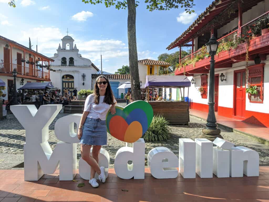 Girl in white t-shirt and jean skirt standing in front of a sign that says "Yo heart Medellin" in Colombia.