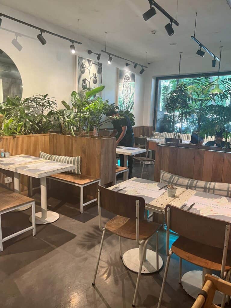 Interior the Kitchen Garden Juhu with empty white tables, wooden benches, and green plants decorating the space.