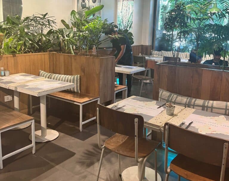 Inside Kitchen Garden Juhu with white tables, wooden chairs and benches with cushions and lots of green plants to decorate the space.