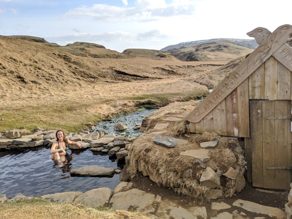 Girl in swimsuit sitting in a natural hot spring in Iceland with wooden changing hut on the left and small rolling hills in the background.