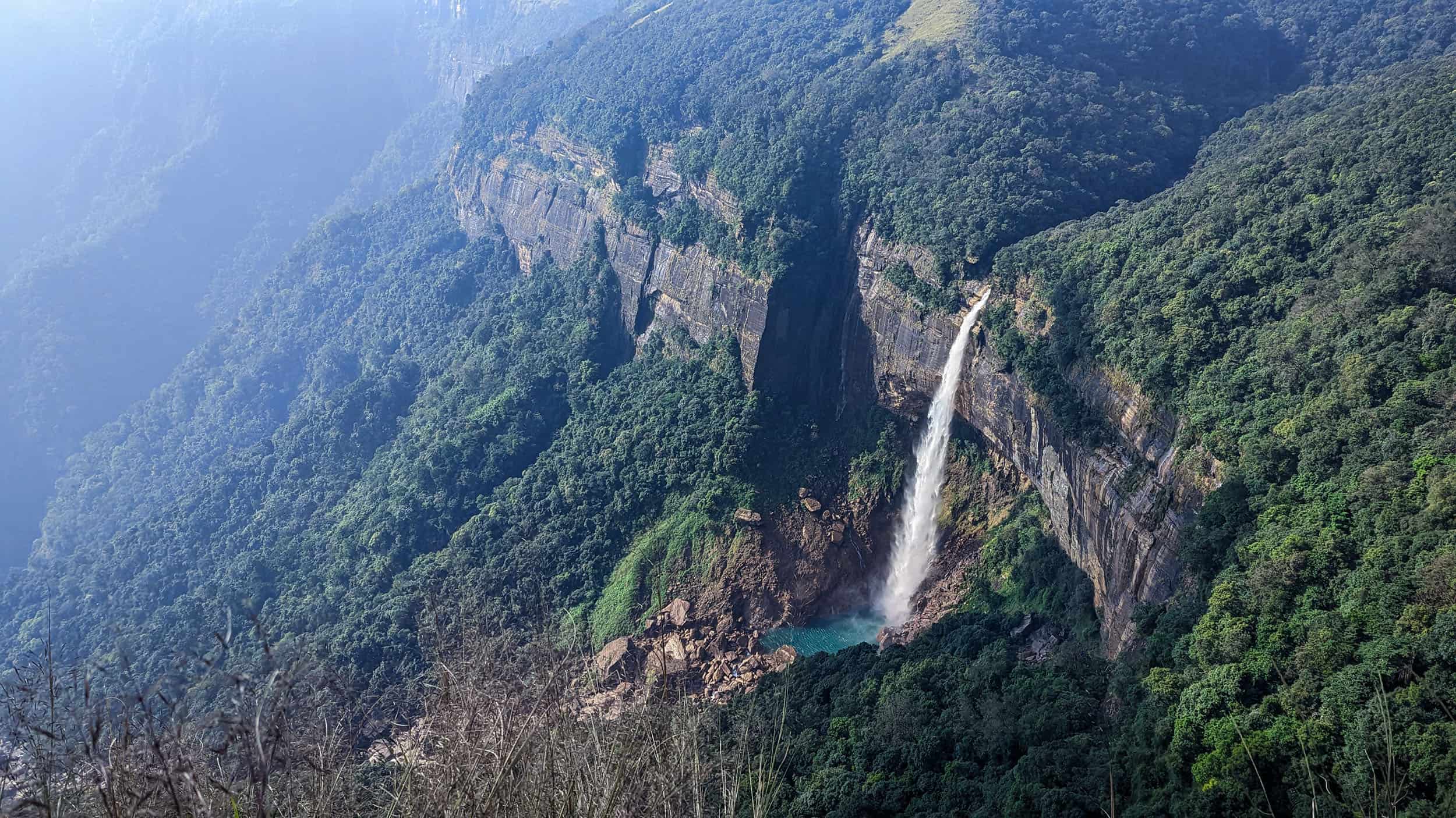 A view of the Nohkalikai falls in Cherrapunji, Meghalaya, India. The picture has been taken from a height and a large column of water is falling down a high cliff with lush green forests. There is an emerald pool of water at the bottom of these high falls and the canyon continues further down as a stream.