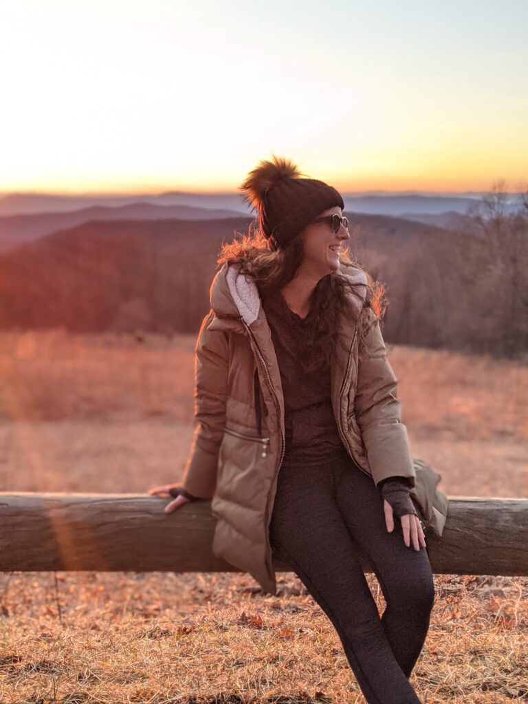 Girl sitting on wooden fence with winter jacket and beanie with the sun setting over the hills in the background.