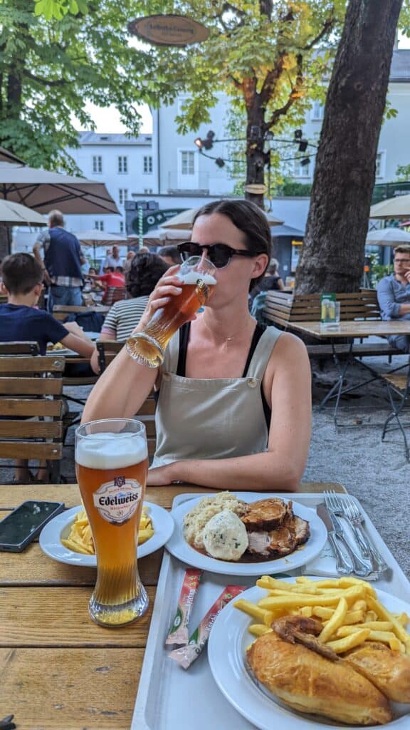 Girl sitting at a picnic table drinking beer at a beer garden in Austria with plates of food in front of her.