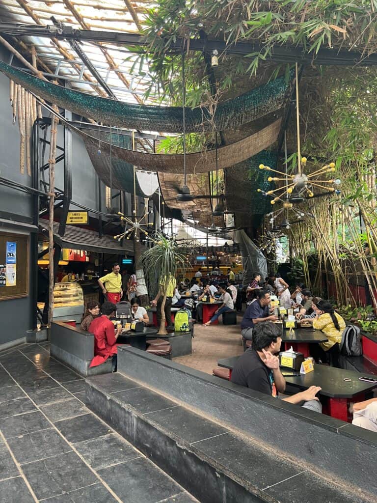 Outdoor cafe next to Prithvi theater in Juhu with people sitting at little black tables and one side with a wall of green bamboo to separate it from the street.