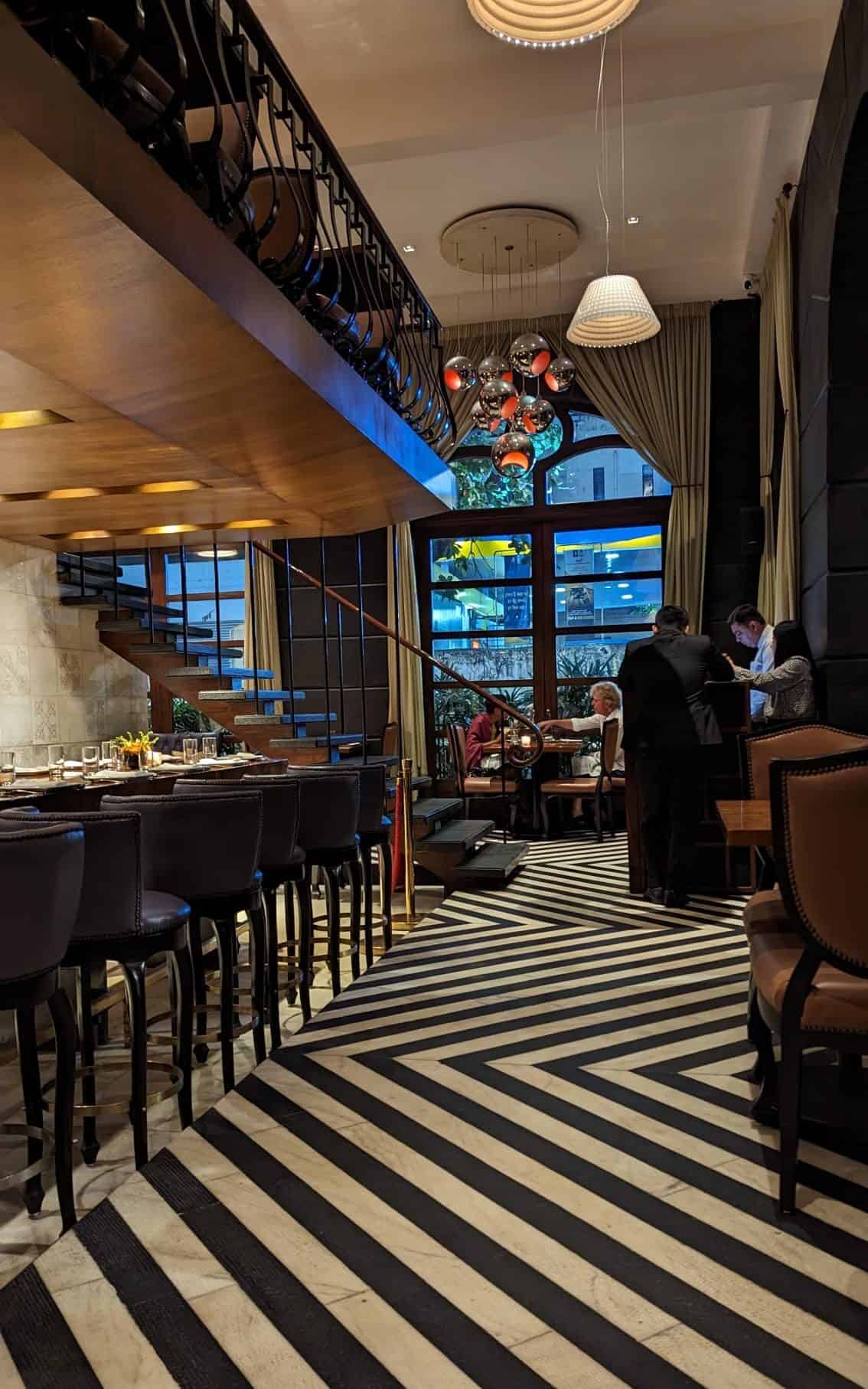 The interior of The Table in South Mumbai, a nice restaurant with a black and white floor, a long bar table, and second floor balcony with more tables.