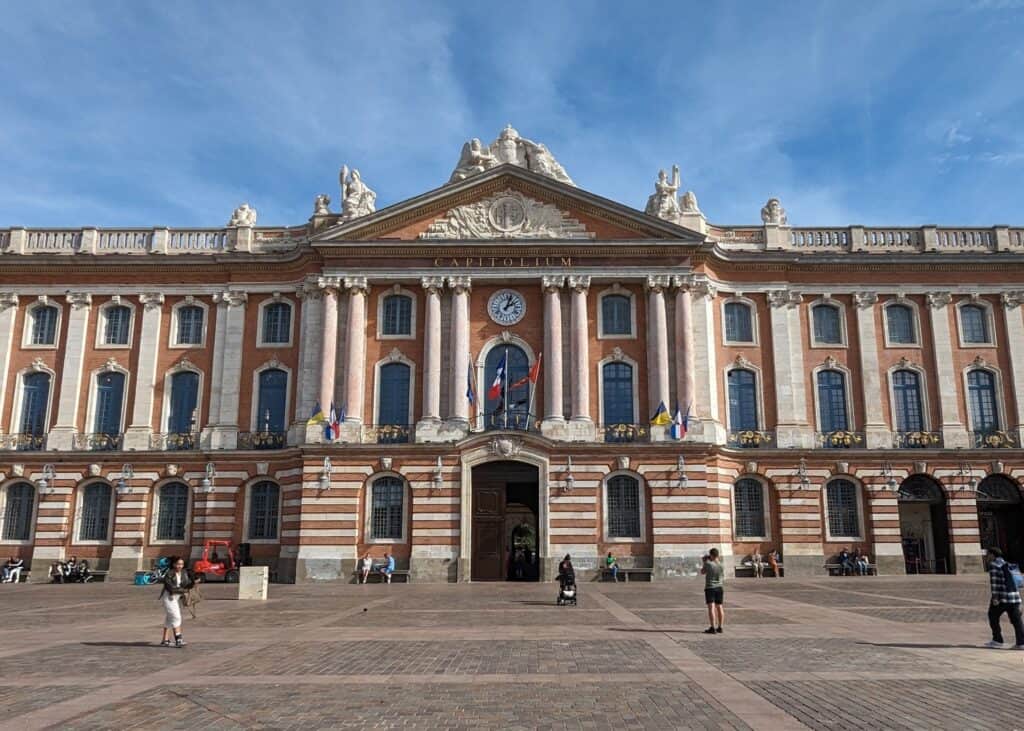 Le Capitole building in Toulouse France on a sunny day. It is a grand building with pink-ish brick and grand white columns.