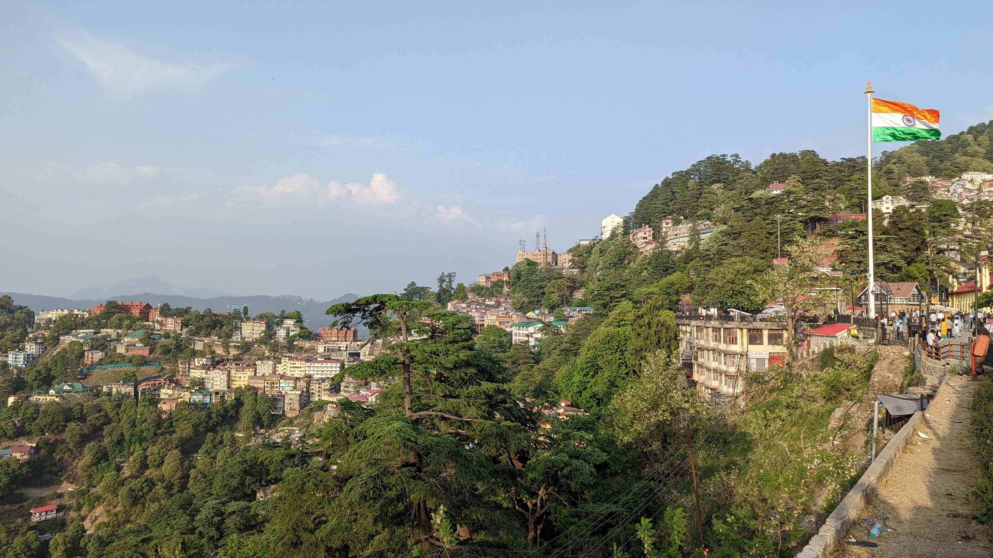 View of Shimla from Mall Road with buildings in the distance and evergreen trees in the hills. An India flag flies in the wind to the right of the view.