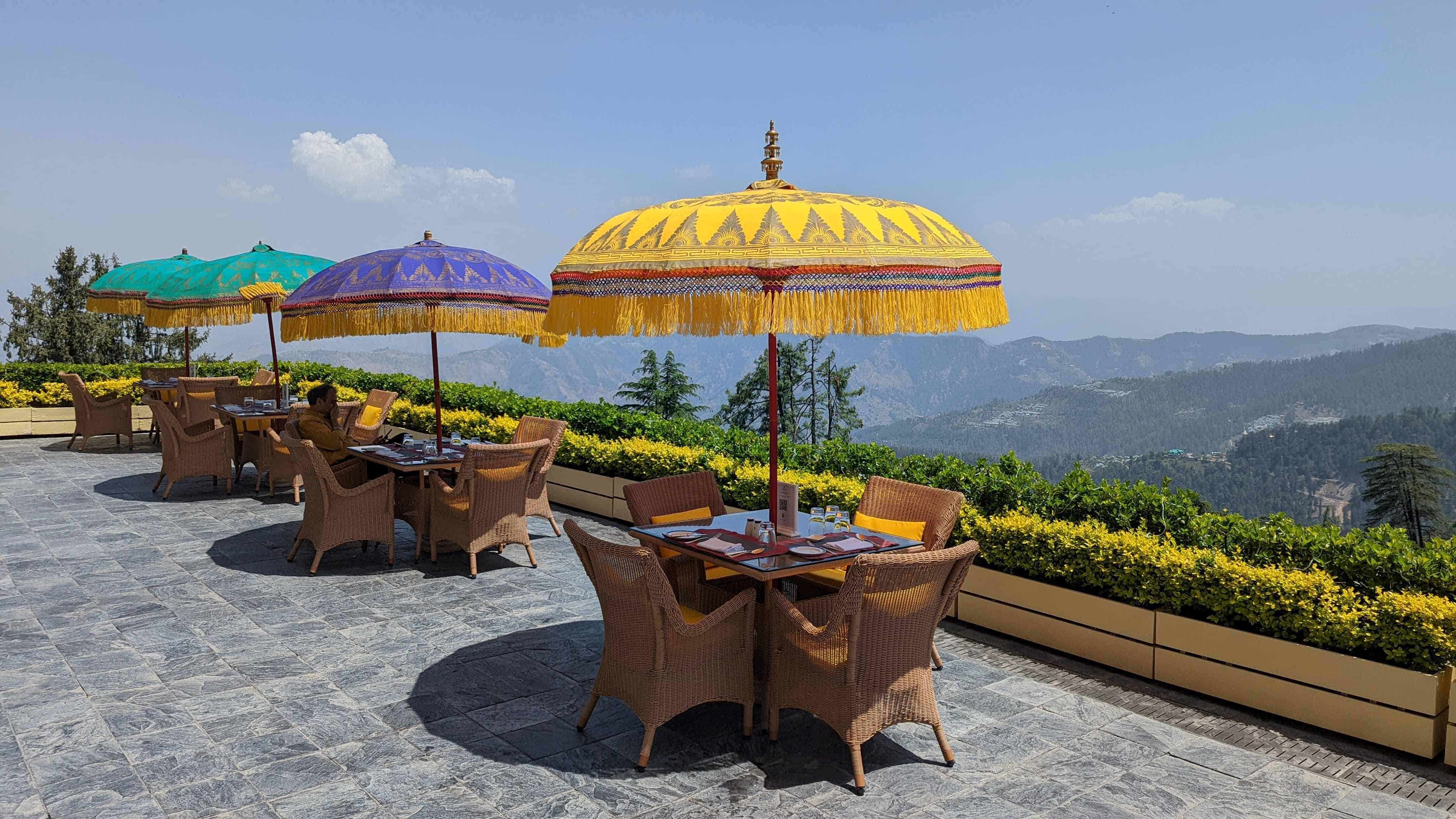 Outdoor deck at Oberoi Wildflower Hall hotel with colorful umbrellas and rattan chairs around empty tables and the mountains in the distance.