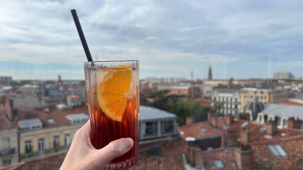 Cocktail with an orange held out in front of cityscape of Toulouse with terracotta colored rooftops in the background.