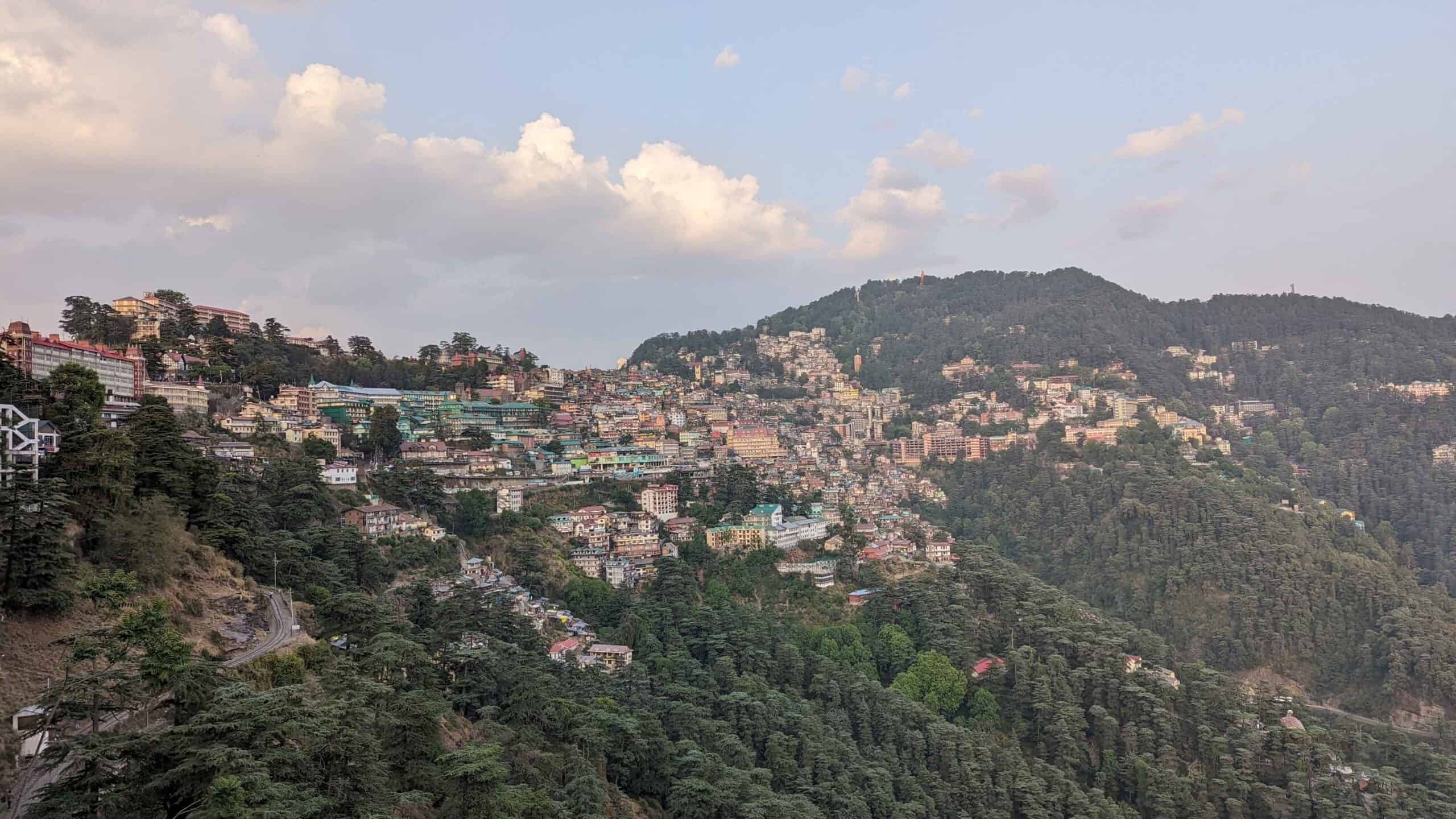 View of Shimla from the distance with colorful buildings interspersed with rolling green hills with evergreen trees.