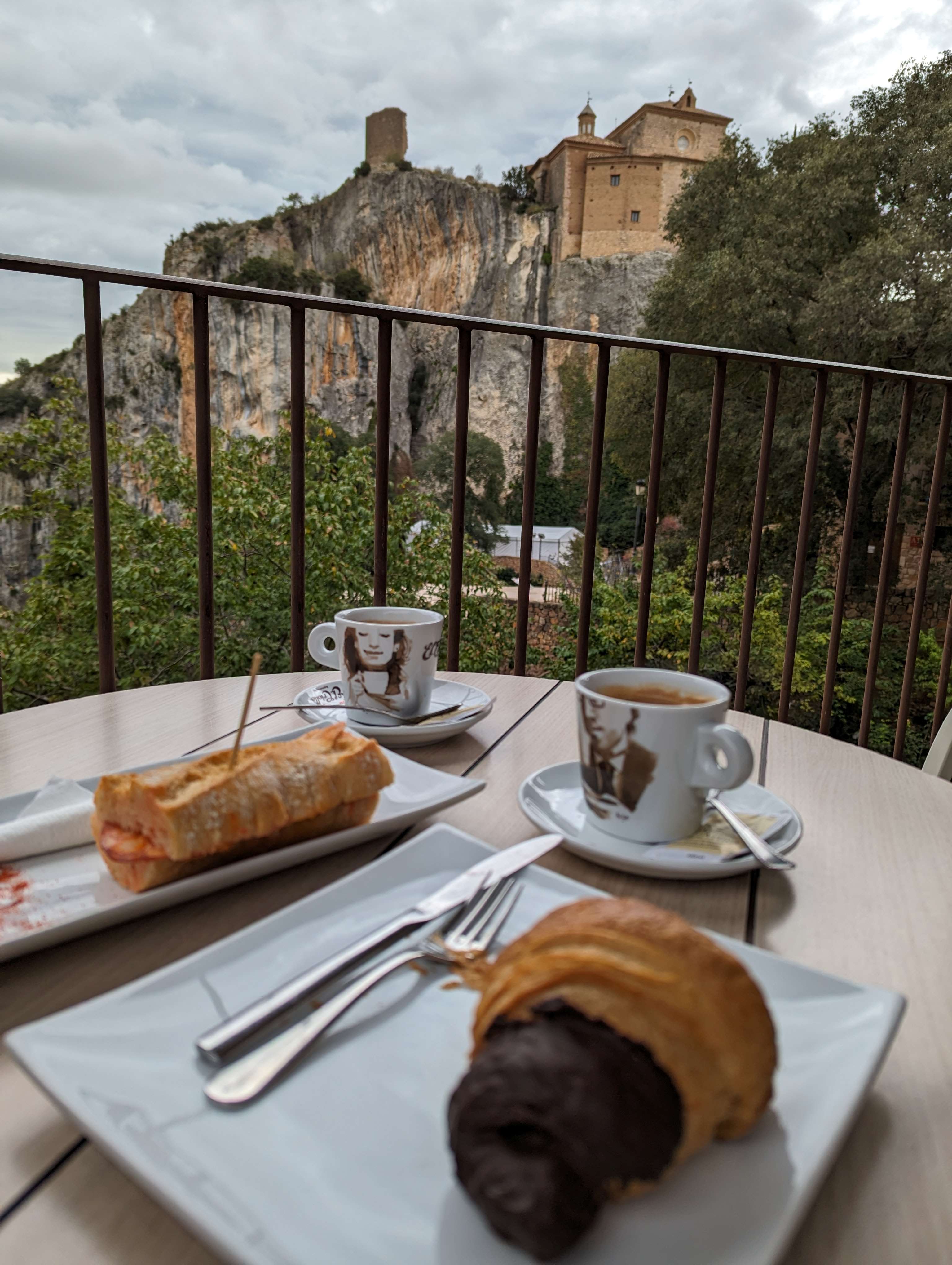 A delicious breakfast spread featuring a chocolate croissant, a ham and cheese baguette, and two cups of coffee, served on a terrace overlooking the dramatic cliffs and historic Collegiate Church of Santa Maria in Alquézar, Spain.