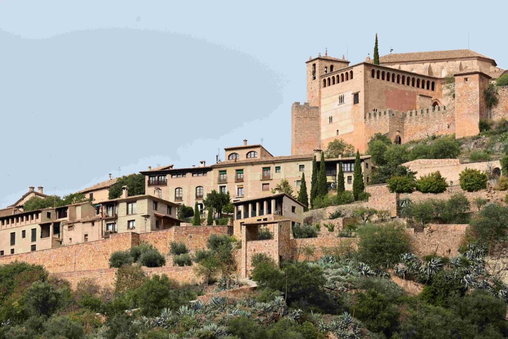 A scenic view of the Collegiate Church of Santa Maria in Alquézar, Spain, perched atop a hill surrounded by well-preserved medieval walls, traditional stone houses, and lush greenery, showcasing the charm of this historic village.