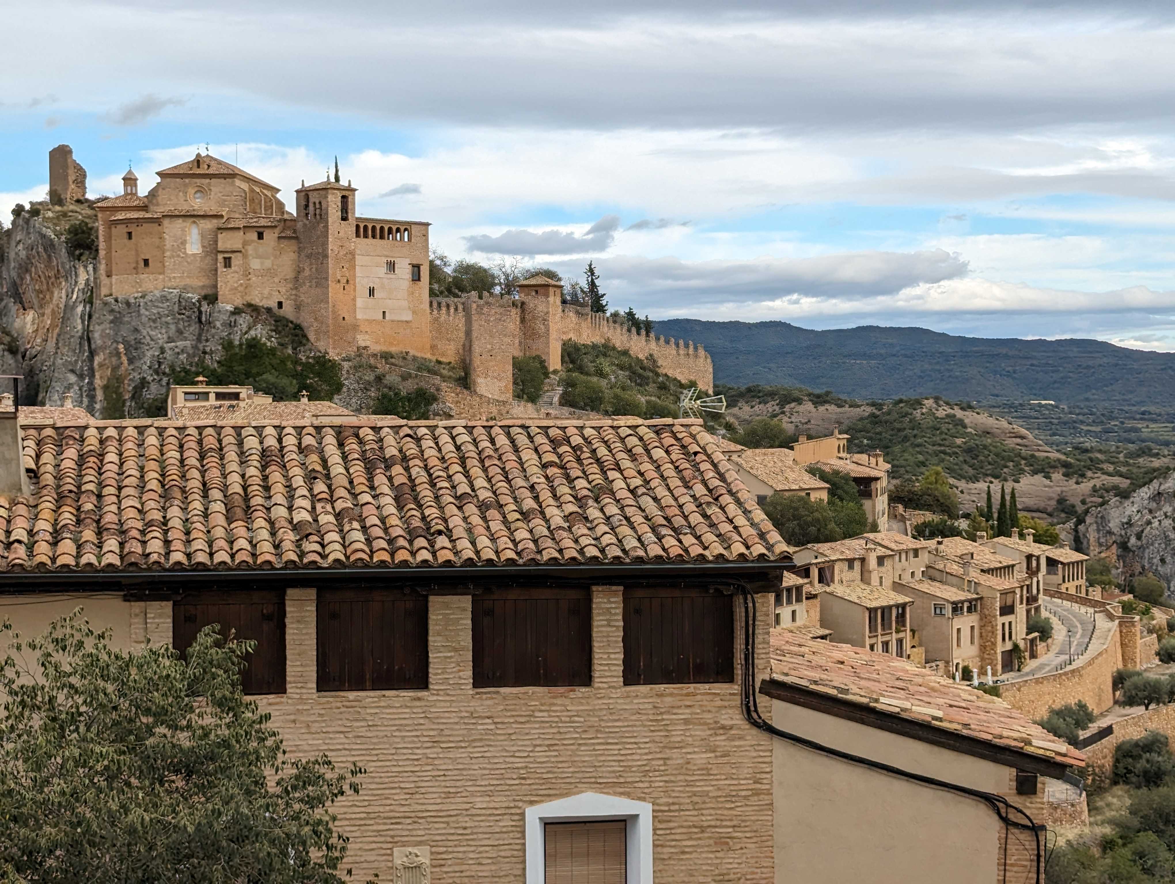 A stunning view from the balcony of Hotel Castillo in Alquézar, Spain, showcasing the historic Collegiate Church of Santa Maria and its surrounding fortified walls, with charming terracotta-roofed houses in the foreground and rolling hills in the distance under a partly cloudy sky.